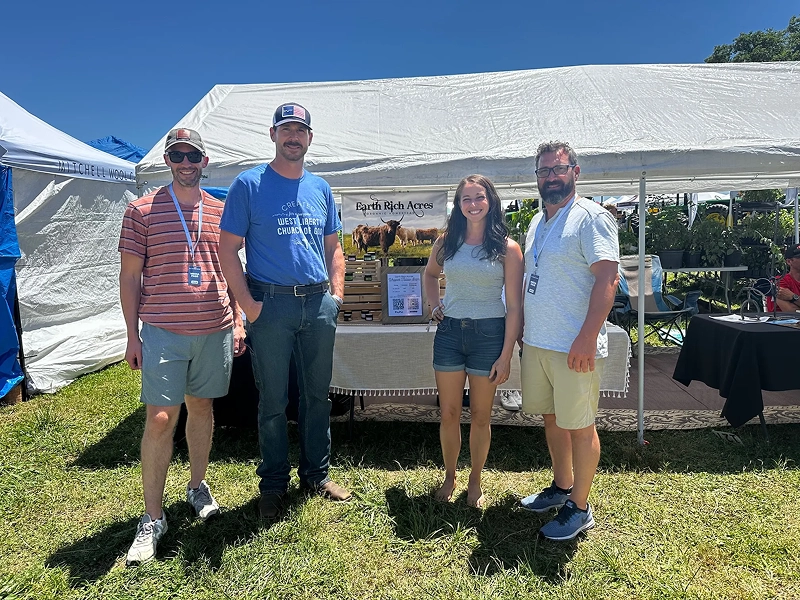 02 Four people standing in front of a white tent at an outdoor event, with a table and display behind them, on a sunny day.