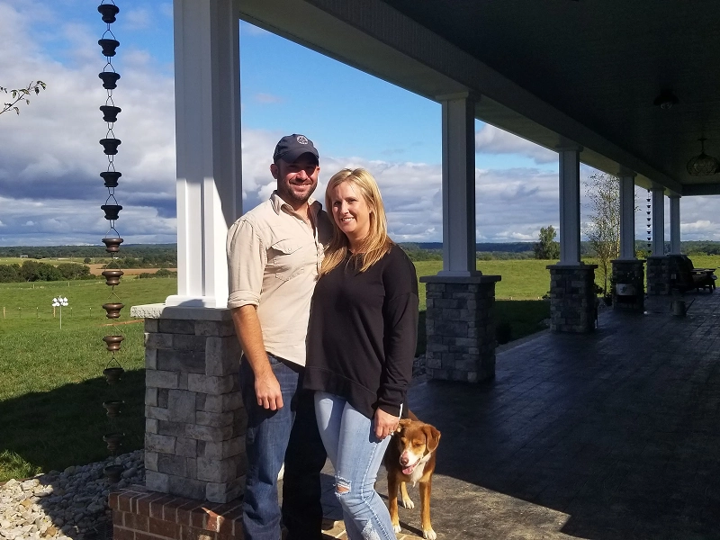 16 A couple poses under a covered patio with a dog. Rolling green fields stretch into the distance under a partly cloudy sky.